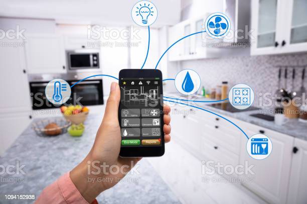 Woman's Hand Using Home Control System On Cellphone With Various Icons In The Kitchen