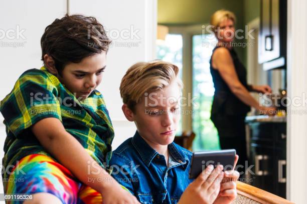 A mother looks on from the kitchen as two pre-teen boys use a smartphone in the living room.  She is out of focus but it is clear to see her sense of discomfort with what they are doing on the internet.