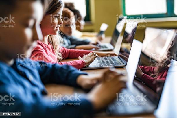 Happy schoolgirl and her classmates e-learning over laptops during a class in the classroom.